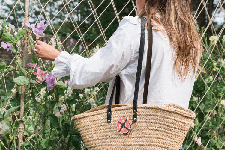 Person holding a woven bag with a floral charm in a garden setting