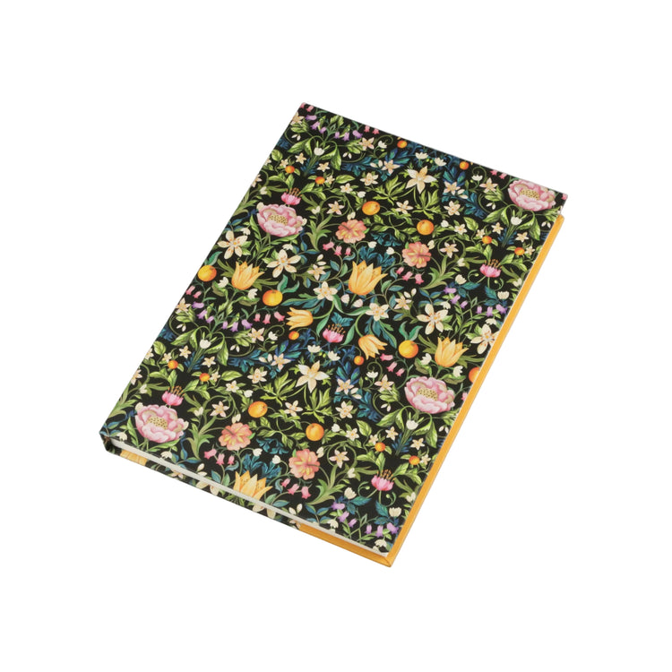 A hardcover notebook with a dense floral pattern on the cover, featuring pink, yellow, and white flowers with green leaves on a dark background, and a yellow-orange spine, photographed on a light gray background.